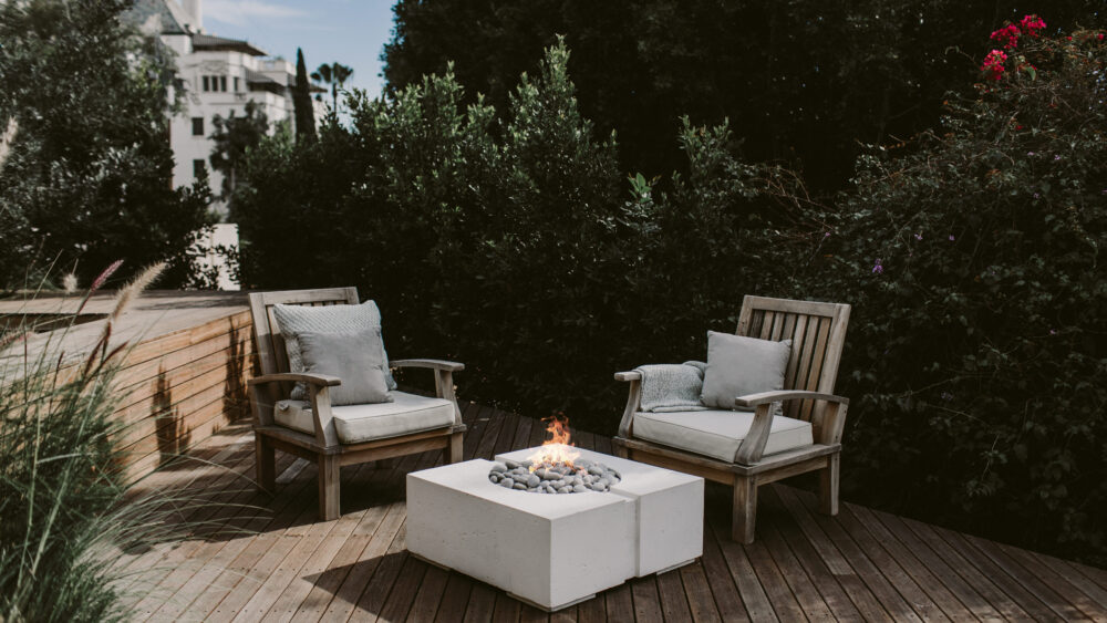 Green garden with a wooden deck in the middle. Two outdoor chairs surround a concrete fire pit.