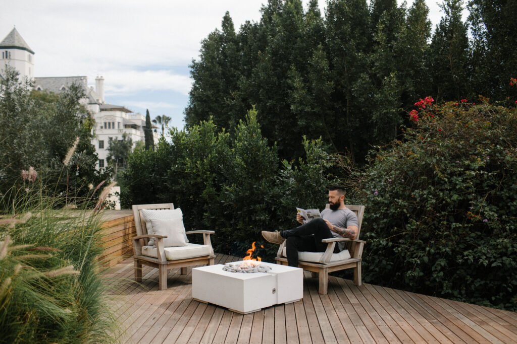 Green garden with a wooden deck in the middle. A man sitting at one of two outdoor chairs, reading a book beside a concrete fire pit.