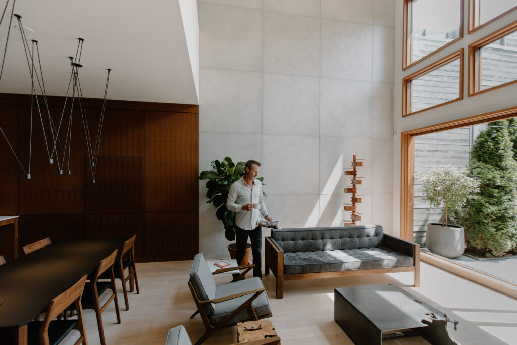 Stylish, modern living space designed with concrete cladding. Middle-aged man walking to a couch with a coffee and magazine. 