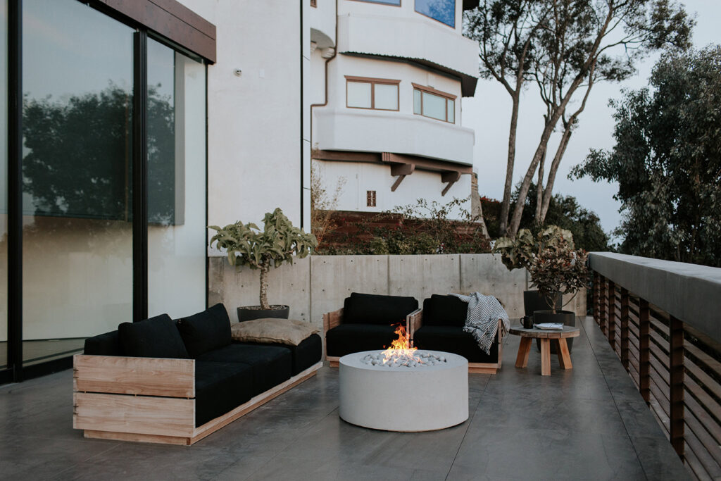 An exterior view showcasing a modern design patio and the back side of a white home. The space features a concrete fire pit, a sitting area, and a city view. Surrounded by greenery and plants. 