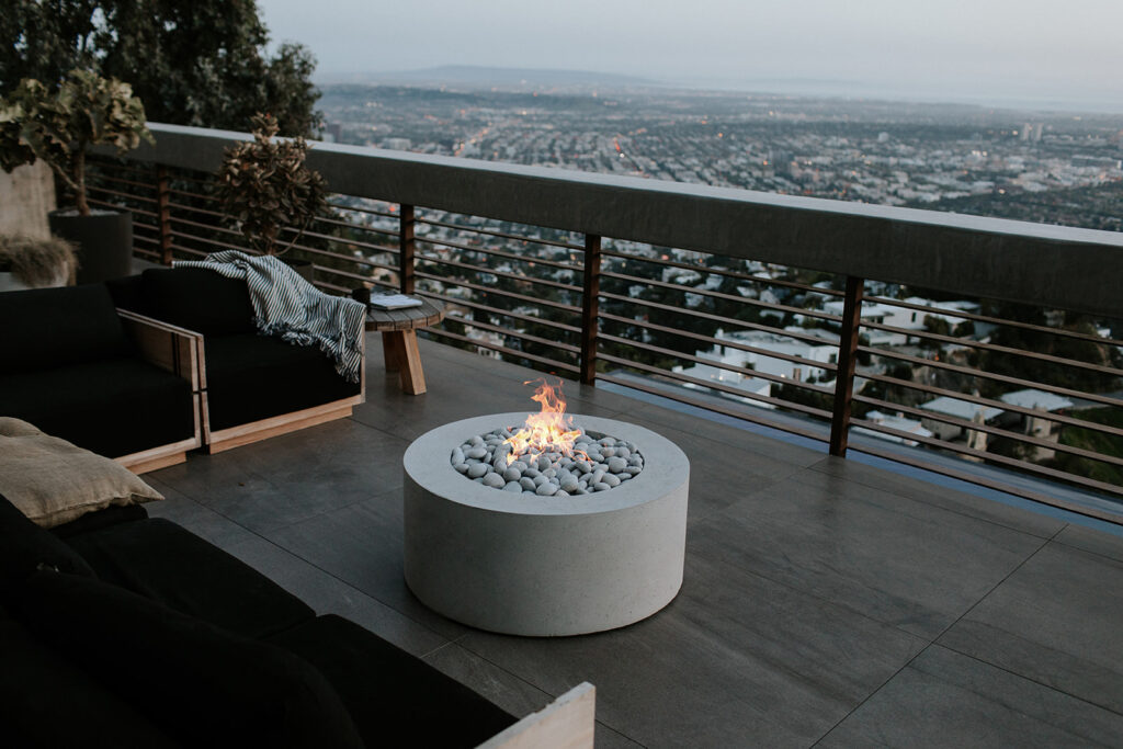 An exterior view showcasing a modern design patio. The space features a concrete fire pit, a sitting area, and a city view. 