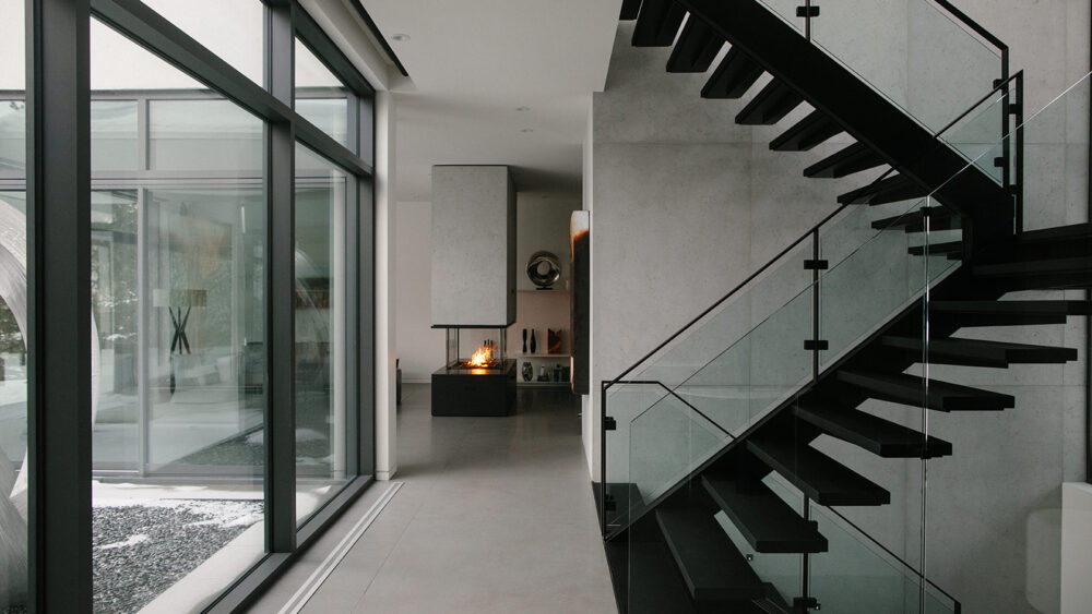 Interior of a modern home, with large windows. Black staircase on the backdrop of a concrete feature wall. Fireplace burning in the back.