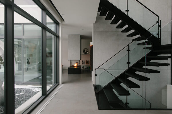 Interior of a modern home, with large windows. Black staircase on the backdrop of a concrete feature wall. Fireplace burning in the back.