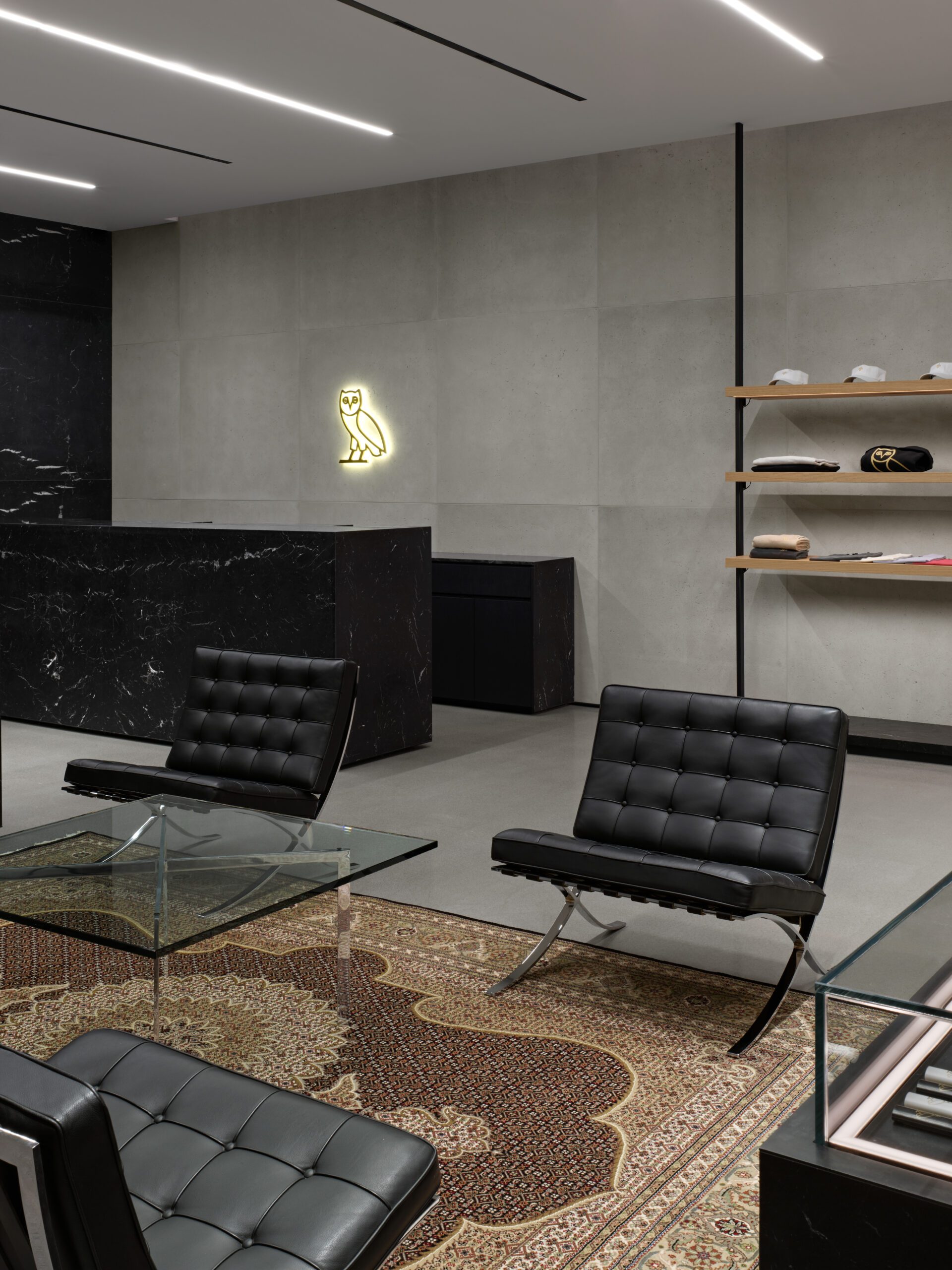 Modern retail space with black leather chairs and glass table, featuring OVO logo, clothing display shelves, and concrete cladding design.