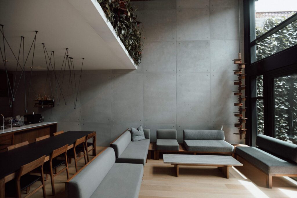 A chic dining and living area with minimalist design elements, characterised by a long dining table with wooden chairs and a sectional sofa. The walls are covered in smooth concrete cladding, complemented by modern, black pendant lights hanging from the ceiling. Large windows allow natural light to illuminate the space, highlighting the clean lines and elegant decor.