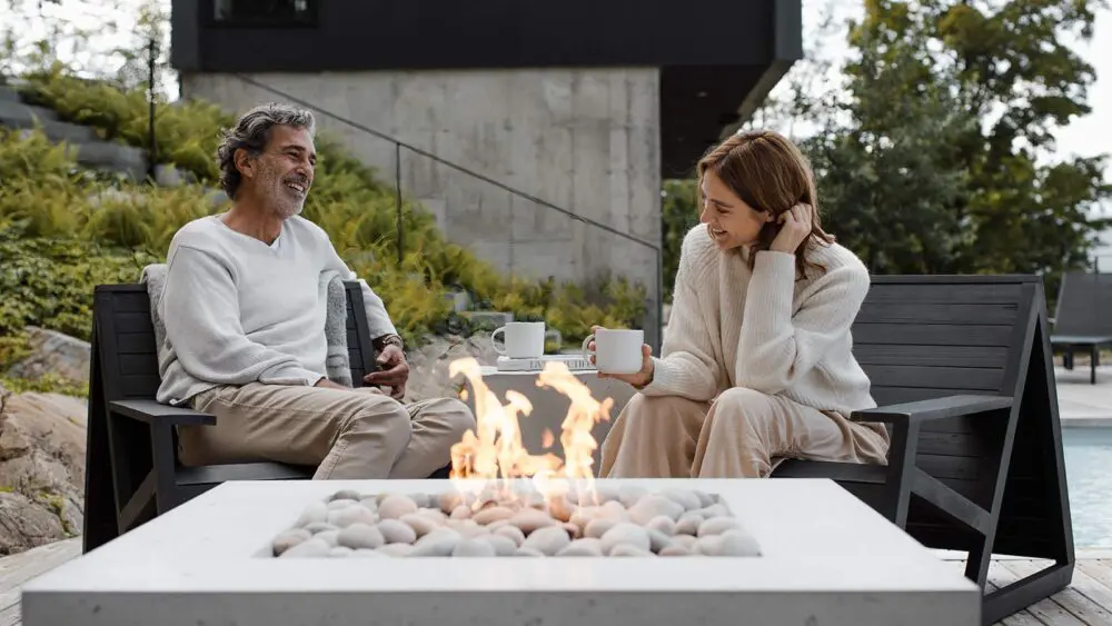 Two people enjoying a cozy moment with hot beverages by a modern concrete outdoor fire pit, surrounded by lush greenery and a contemporary house in the background.