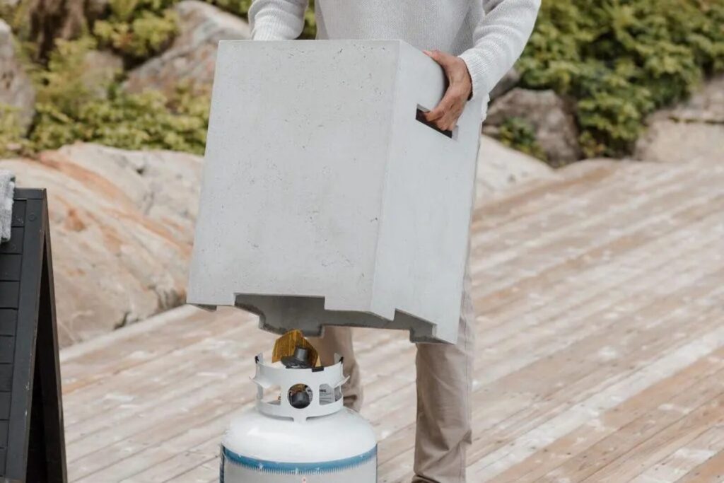 Man placing a concrete tank cover over a propane tank on a wooden deck with lush greenery in the background. 