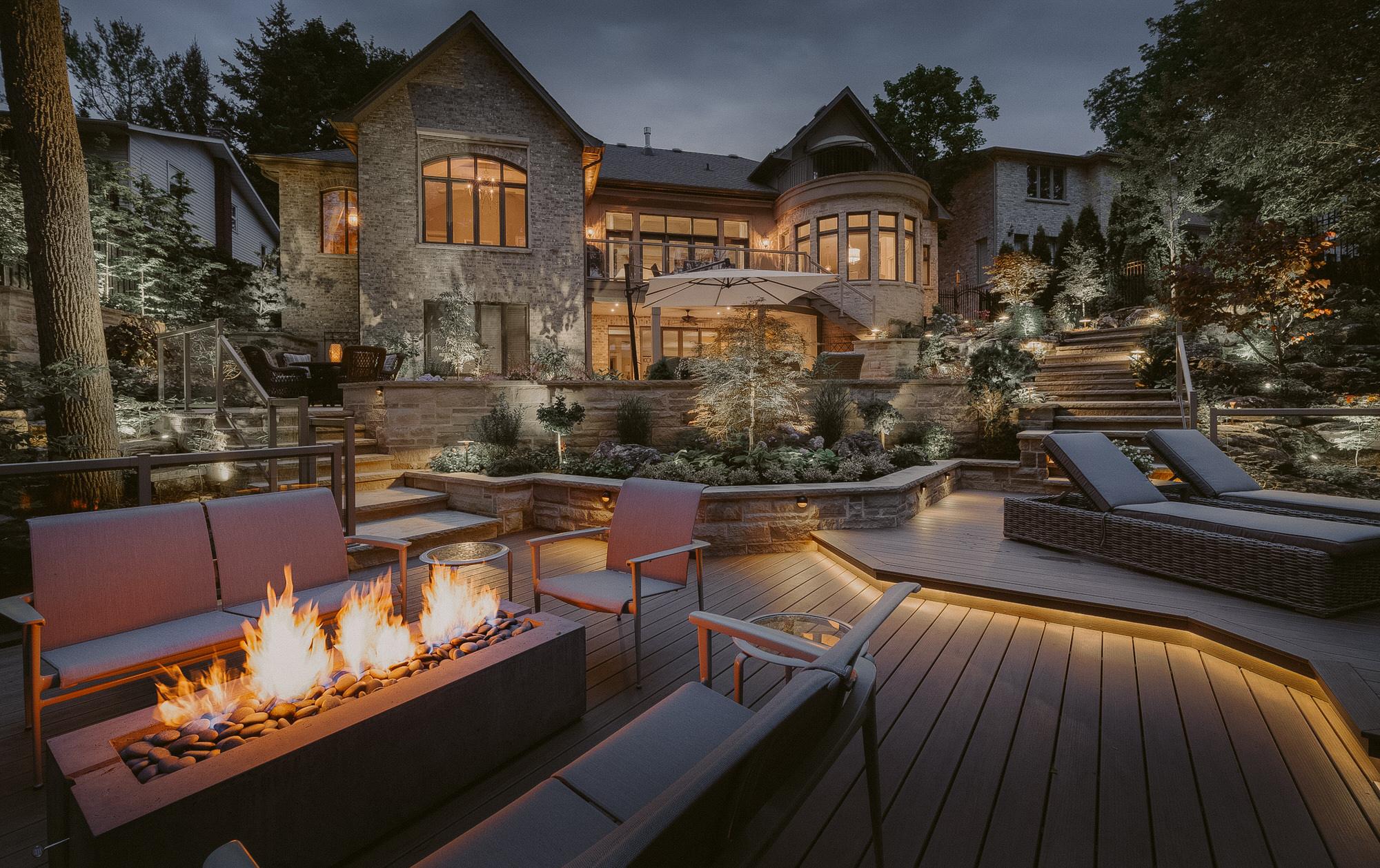 A Dekko Avera concrete fire pit in the middle of gray patio furniture in front of a stone two-story home with an abundance of foliage in the yard.