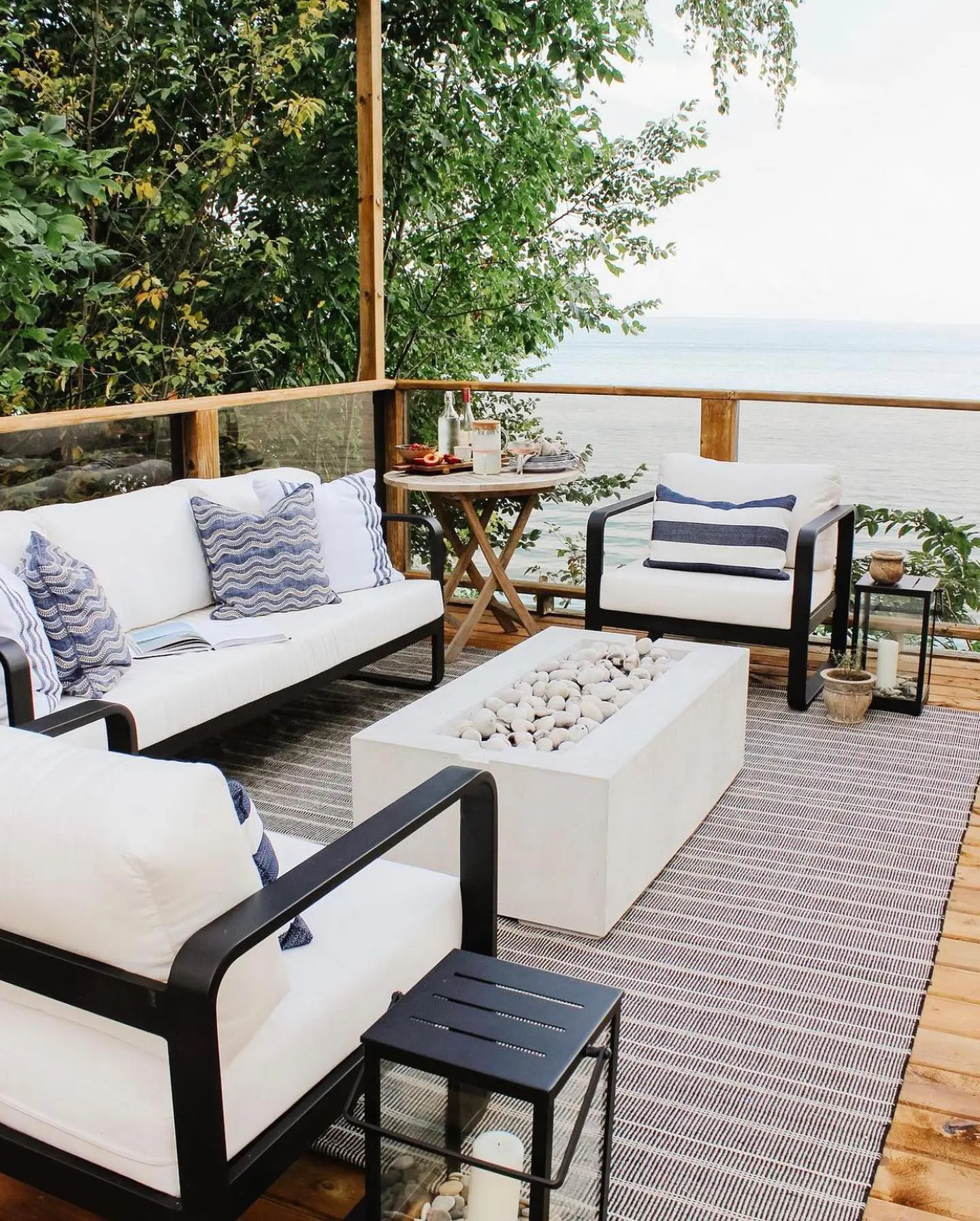 Two black-framed chairs and a couch with cream-colored cushions and navy and cream-colored pillows are arranged around a cream-colored Avera fire pit. 