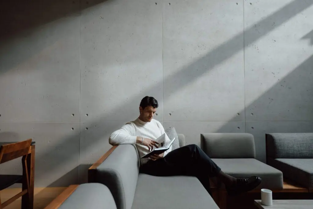 A chic living area with minimalist design elements, characterised by a long dining table with wooden chairs and a sectional sofa. The walls are covered in smooth concrete cladding, complemented by modern, black pendant lights hanging from the ceiling. Large windows allow natural light to illuminate the space, highlighting the clean lines and elegant decor. A young man is sitting on the sectional couch reading a book. 