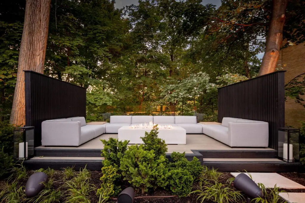 Stylish outdoor seating area featuring a rectangular concrete fire pit, surrounded by modern grey sectional sofas. The space is framed by sleek black privacy walls and lush greenery, creating a cozy, nature-inspired retreat.