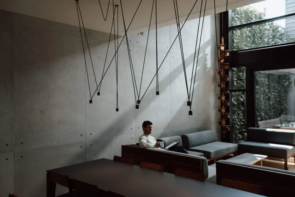 A stylish living room featuring a man sitting on a gray sectional sofa with a book. The space has high concrete walls, large windows with an outdoor view, and unique pendant lights with black wires forming geometric shapes hanging from the ceiling. The room exudes a cozy yet modern ambiance.