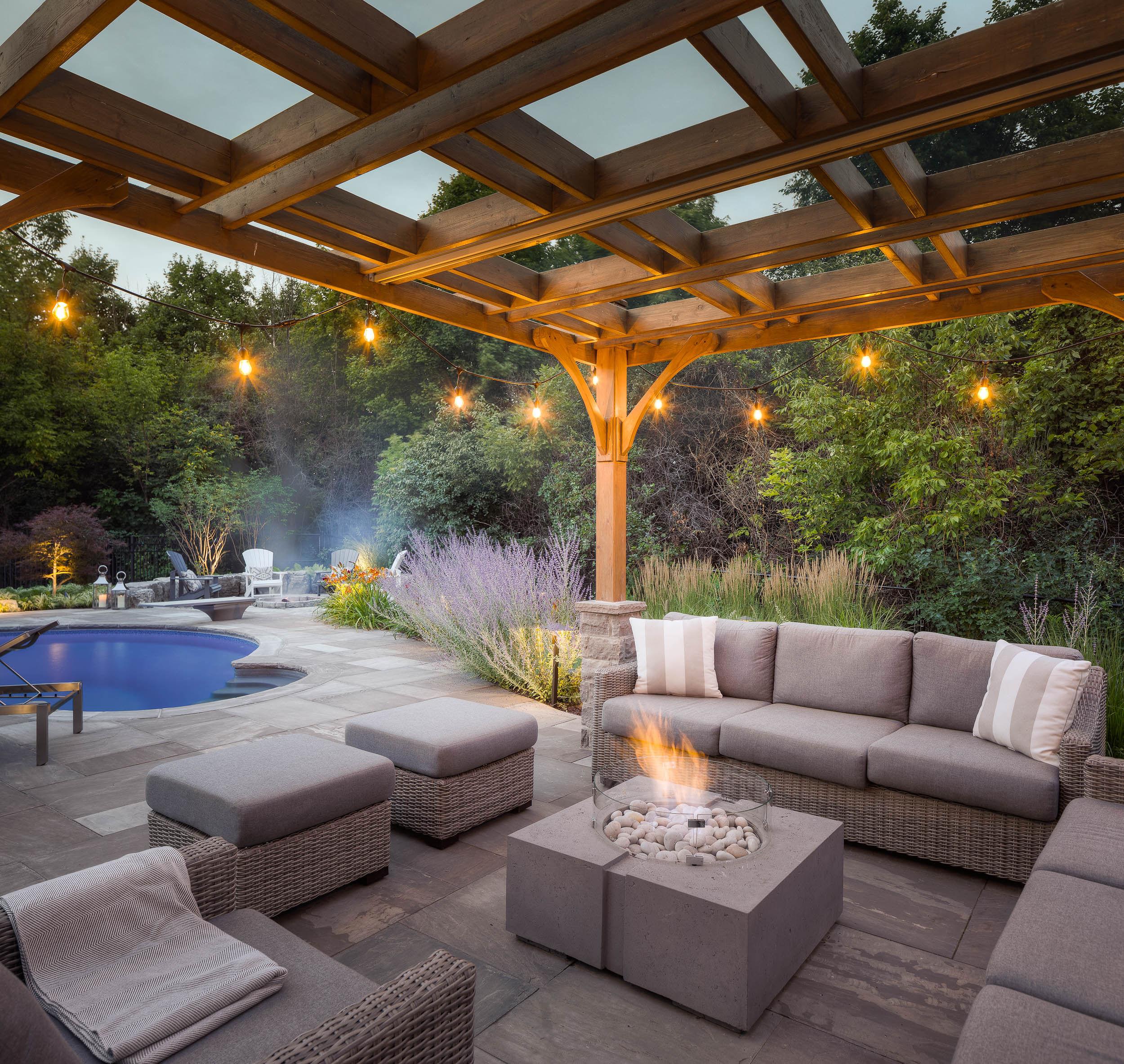A square Dekko lightweight concrete fire pit surrounded by patio furniture under a pergola next to a pool and a lot of landscaping plants.